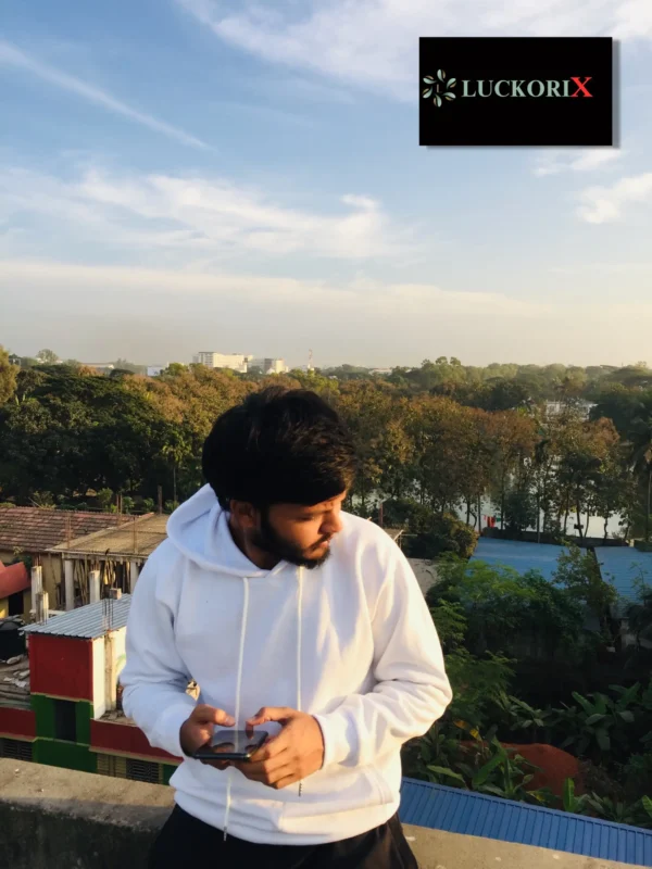 Man wearing a LuckoriX white hoodie outdoors, holding a phone, with greenery and blue sky in the background.